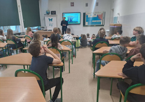 Uczniowie klas czwartych siedzący przed prowadzącym zajęcia na temat burz i innych zjawisk meteorologicznych. W tle ekran na którym wyświetlana jest prezentacja na omawiany temat.
