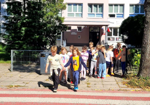Grupa uczniów stojąca przed jezdnią parami, w tle budynek szkoły .