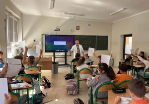 Sala lekcyjna, przy tablicy nauczyciel, przed nim w ławkach siedzą dzieci z podniesionymi kartkami.