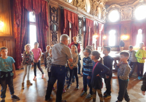 Grupa dziec,i stojąca w pieknych wnętrznach Pałacu Poznańskiego, słuchająca przewodnika.