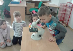 Troje przedszkolakwó siedzi na podłodze, jeden z chłopców ogląda preparaty pod mikroskopem.