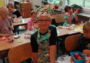 Chłopiec w kolorowym fartuszku i czapce kucharza przygotowujący kanapiki, w tle inne dzieci przy pracy.
