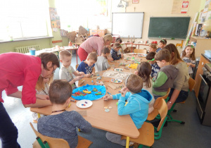 Przedszkolaki siedzące wokół stolika z materiałami plastycznymi, próbujące wykonać zaprezentowane im prace.